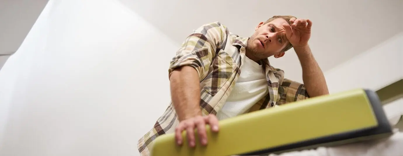 An image of a weary, muscular man trying to close his suitcase with clothes, sweating and wiping his forehead after.